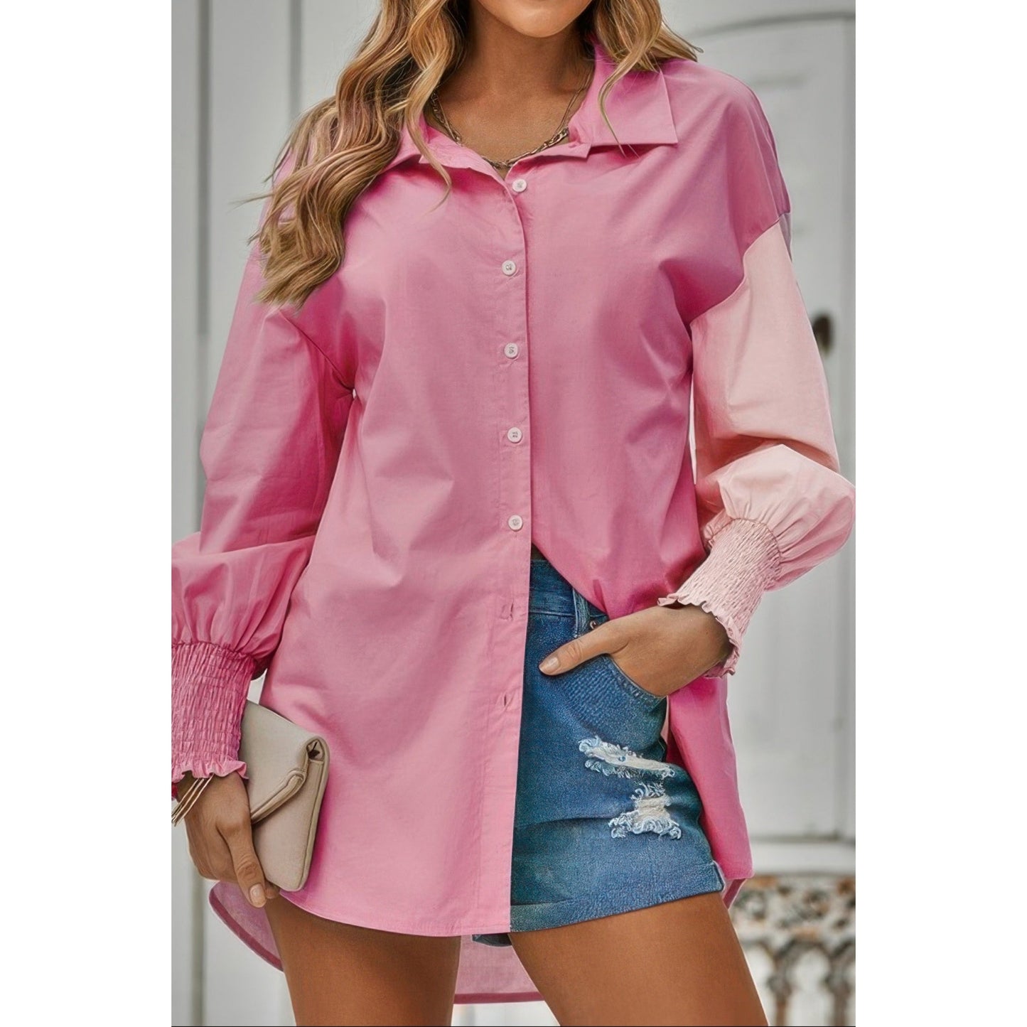 A woman wearing a pink long-sleeve button-up shirt with contrasting light pink sleeves and ruffled cuffs, paired with distressed denim shorts and holding a beige clutch.
