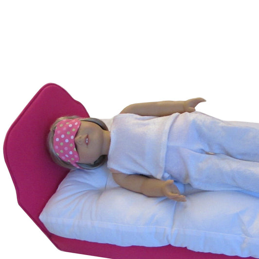 An 18-inch doll wearing a polka dot sleep mask while lying on a pink doll bed.