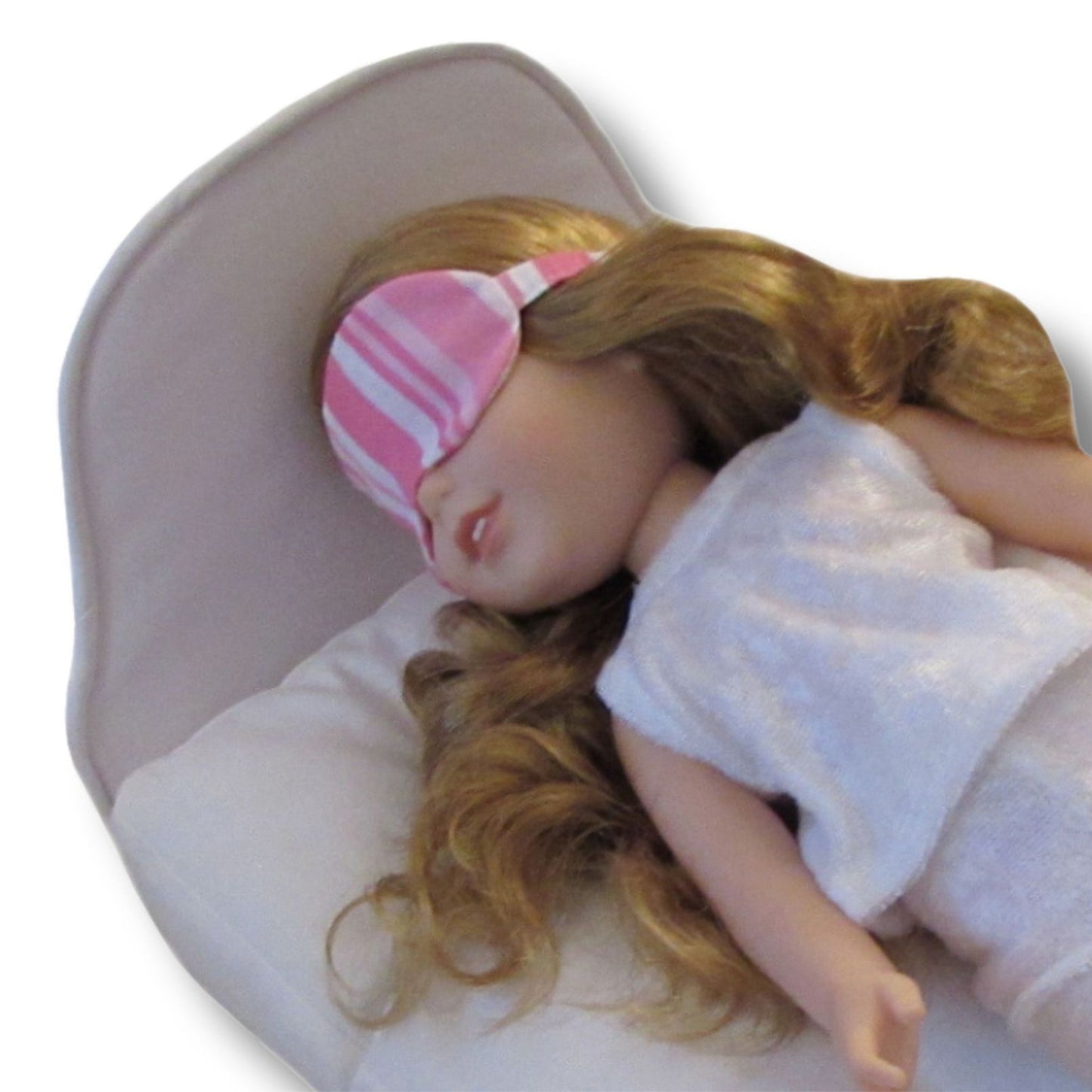 A doll wearing a pink striped eye mask and white pajamas while lying on a white doll bed.
