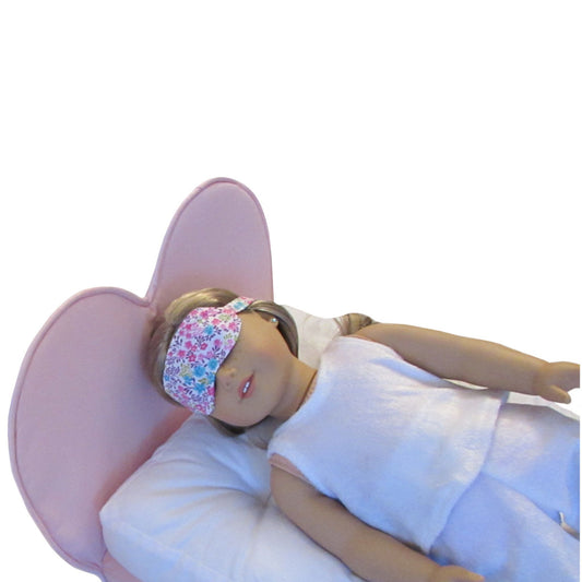 Doll wearing a floral eye mask and white outfit on a pink chair with a white background