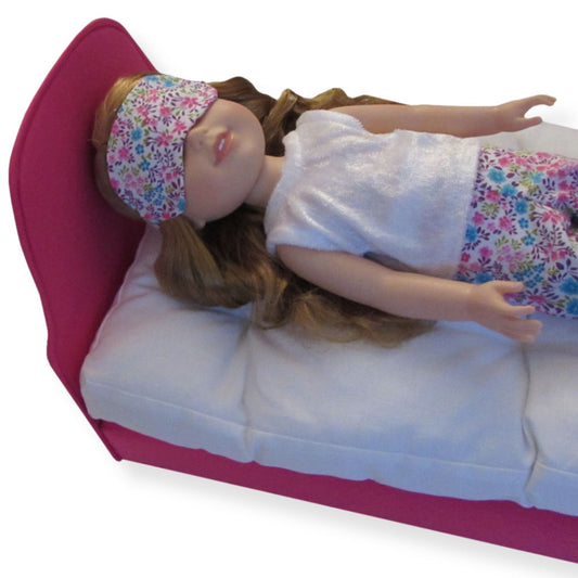 A 14.5-inch doll wearing a pink turquoise floral sleep mask while lying on a pink bed.
