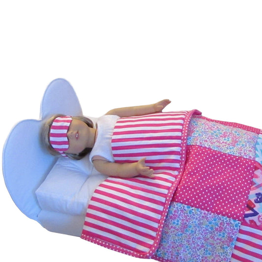 A doll wearing a pink and white striped sleep mask while lying underneath a patchwork quilt.