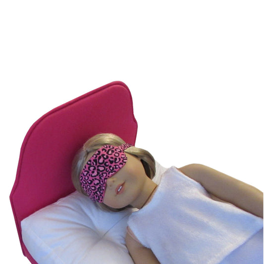An 18-inch doll wearing a pink cheetah print sleep mask while lying on a pink doll bed.