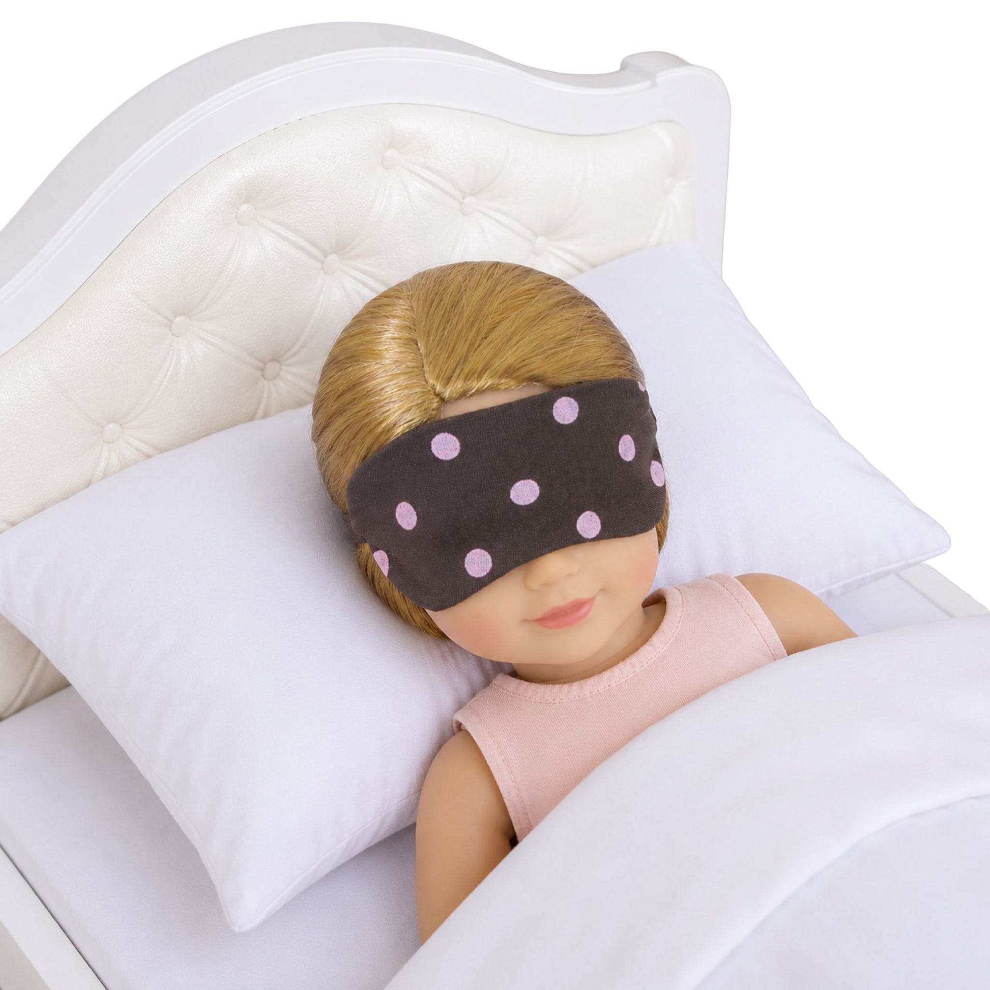 A 14.5-inch doll is wearing a brown sleep mask with pink polka dots, lying in a bed.