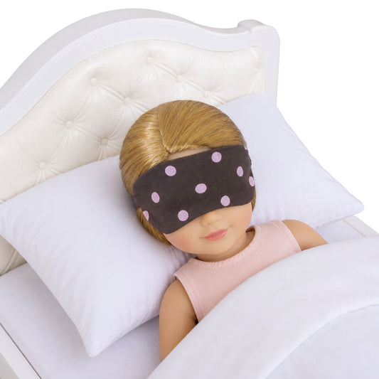 A 14.5-inch doll is wearing a brown sleep mask with pink polka dots, lying in a bed.