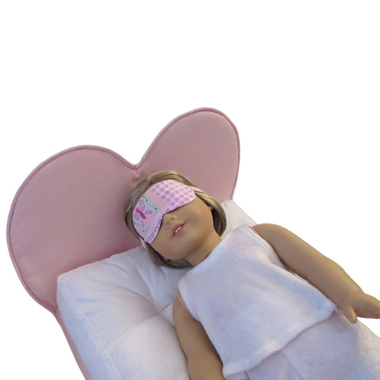 An 18-inch doll wearing a pink patchwork sleep mask while lying on a pink heart doll bed.