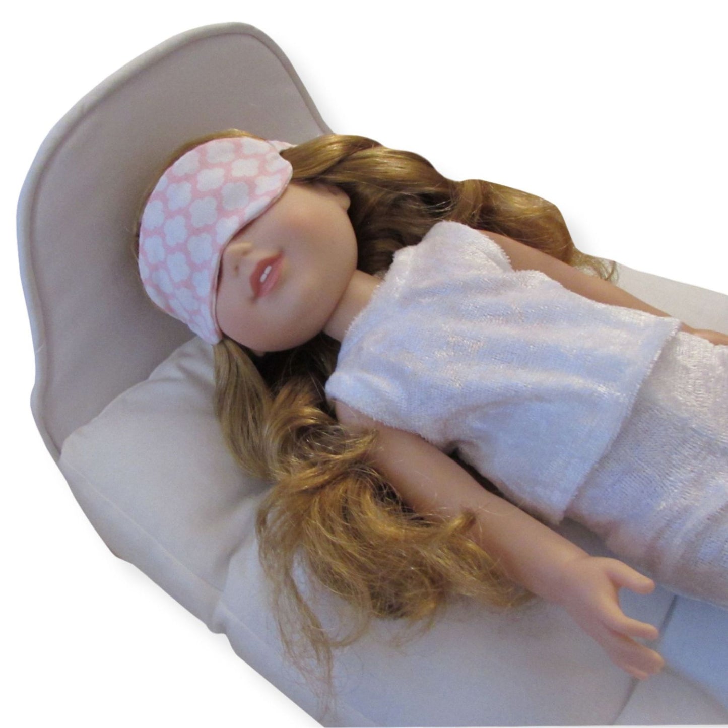 A 14.5-inch doll wearing a pink print sleep mask while lying on a white doll bed.