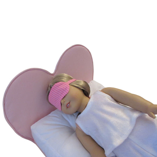 An 18-inch doll is wearing a pink striped sleep mask while lying on a pink heart doll bed.