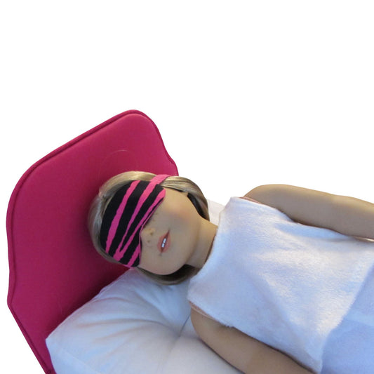 An 18-inch doll wearing a pink zebra print sleep mask while lying on a pink bed.