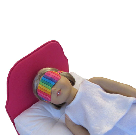An 18-inch doll wearing a rainbow print sleep mask while lying on a pink doll bed.