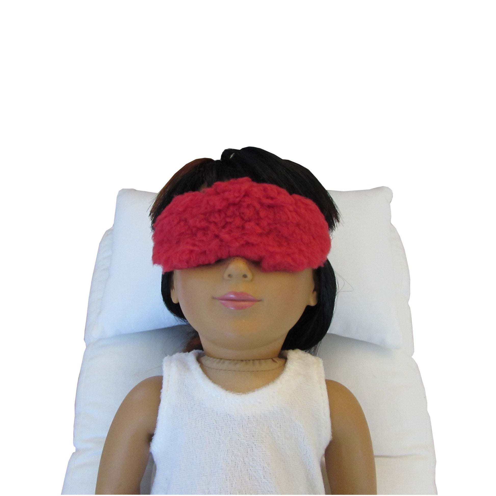 An 18-inch doll is wearing a red faux sherpa fur sleep mask while lying on a white doll pillow and a white doll mattress.
