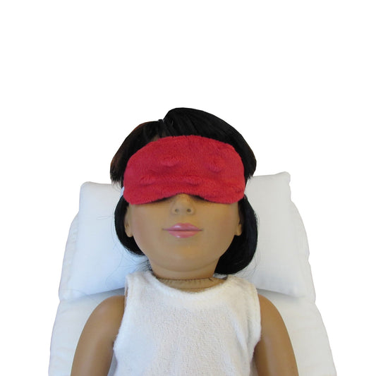 An 18-inch doll wearing a red minky sleep mask while lying on a white doll pillow and a white doll mattress.