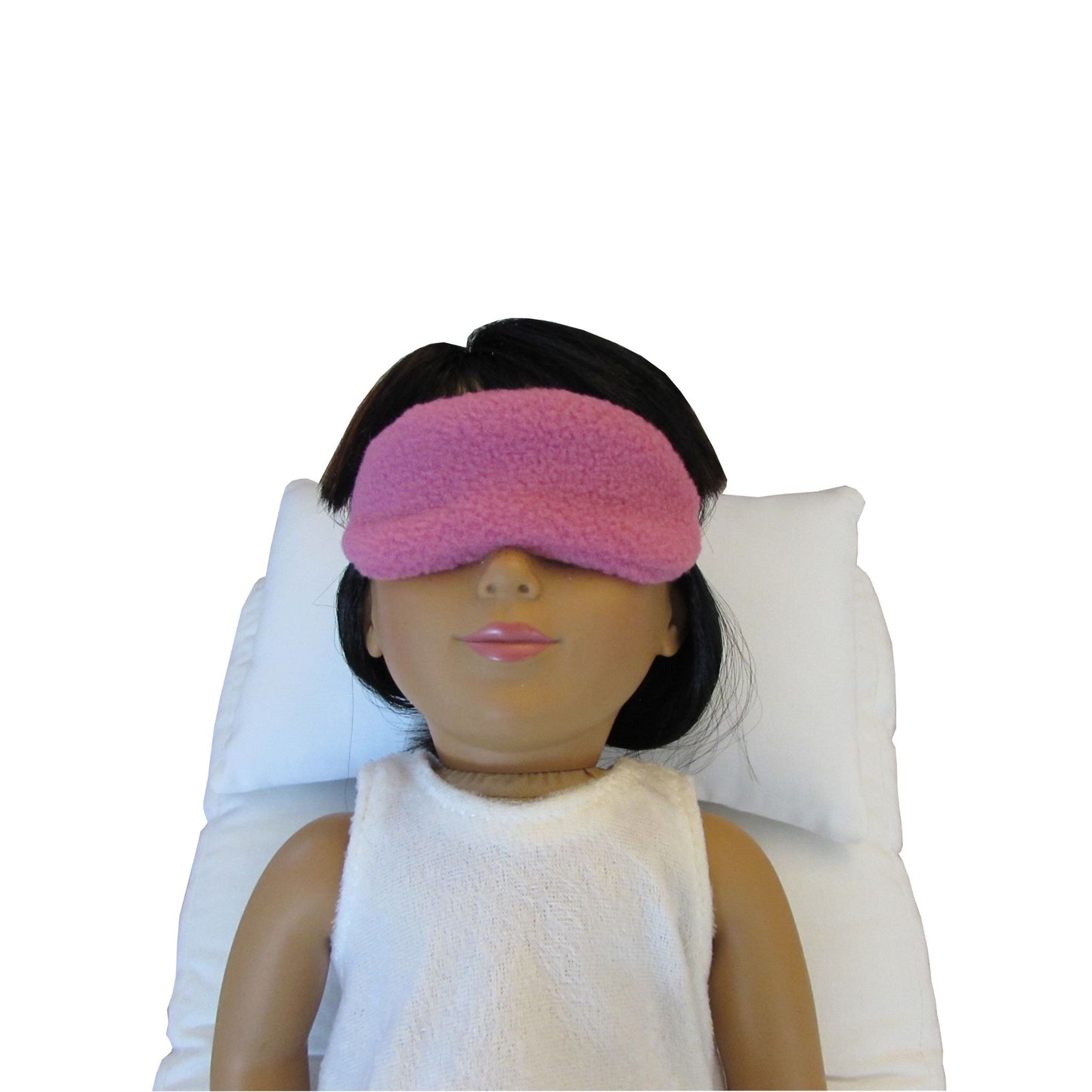 An 18-inch doll is wearing a Rose Anti-Pill Fleece sleep mask while lying on a white doll pillow and a white doll mattress.