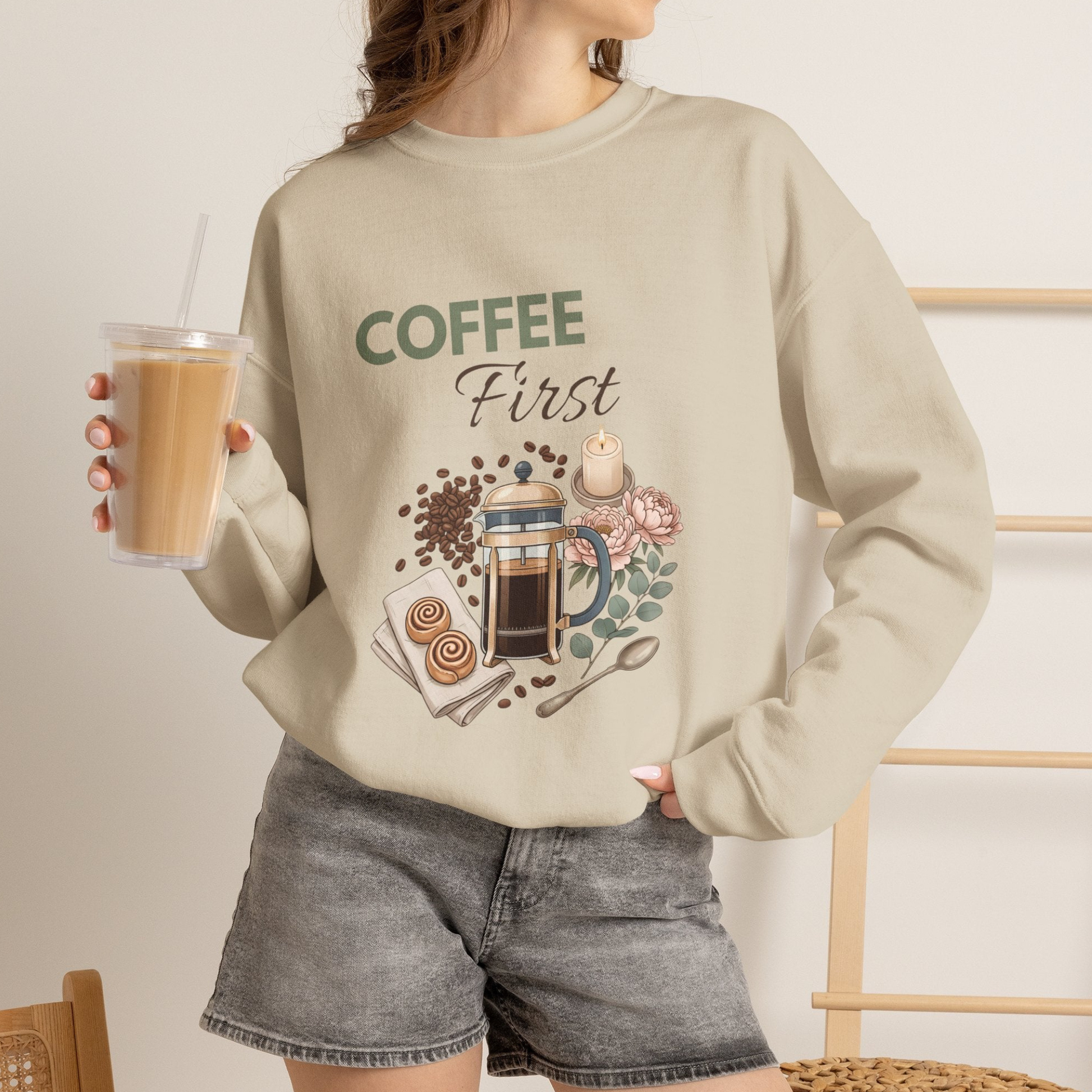 A woman is wearing a sand-colored crewneck sweatshirt with 'COFFEE First' on the front and is holding an iced coffee.