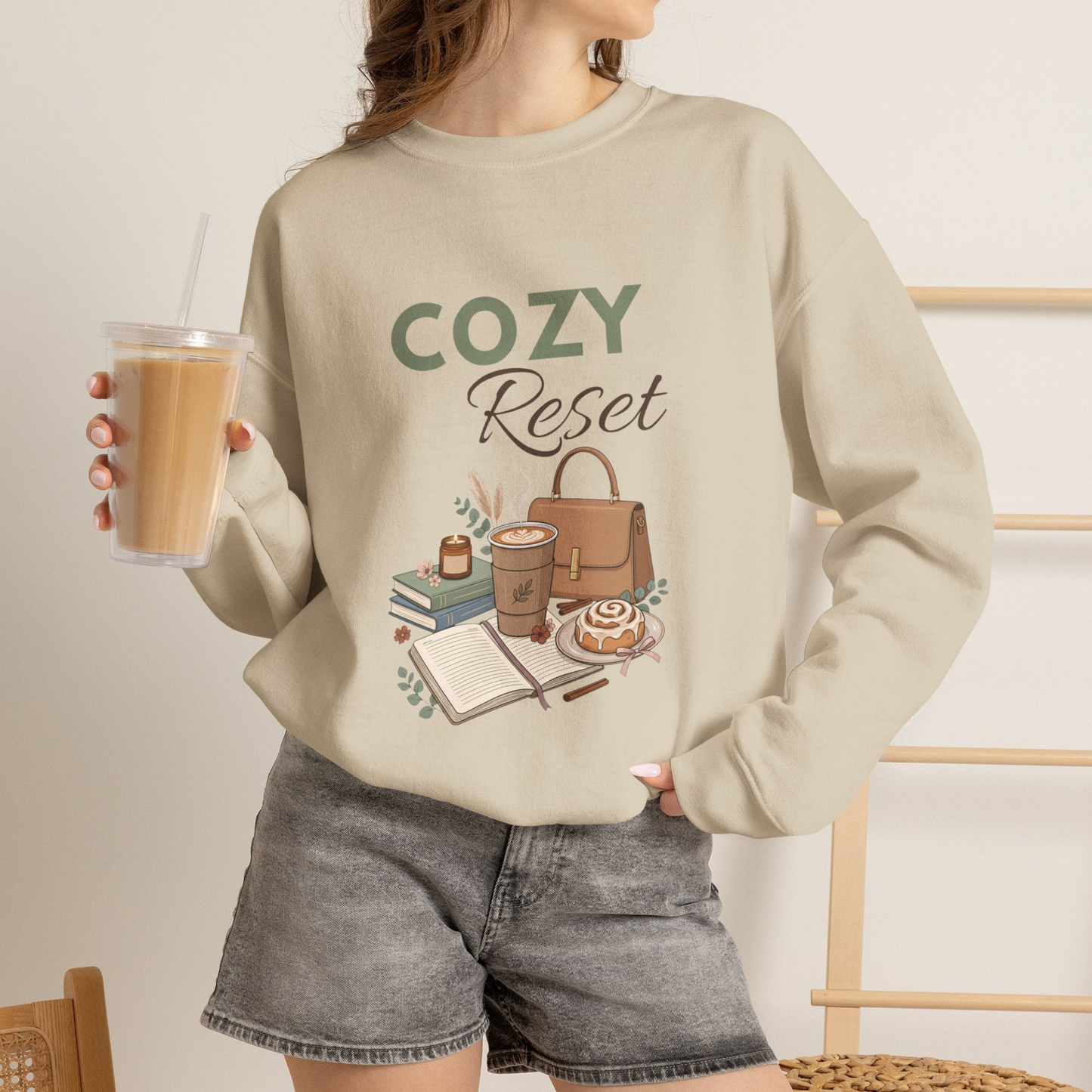 A woman is wearing a sand-colored sweatshirt with 'COZY Reset' on the front and is holding an iced coffee.