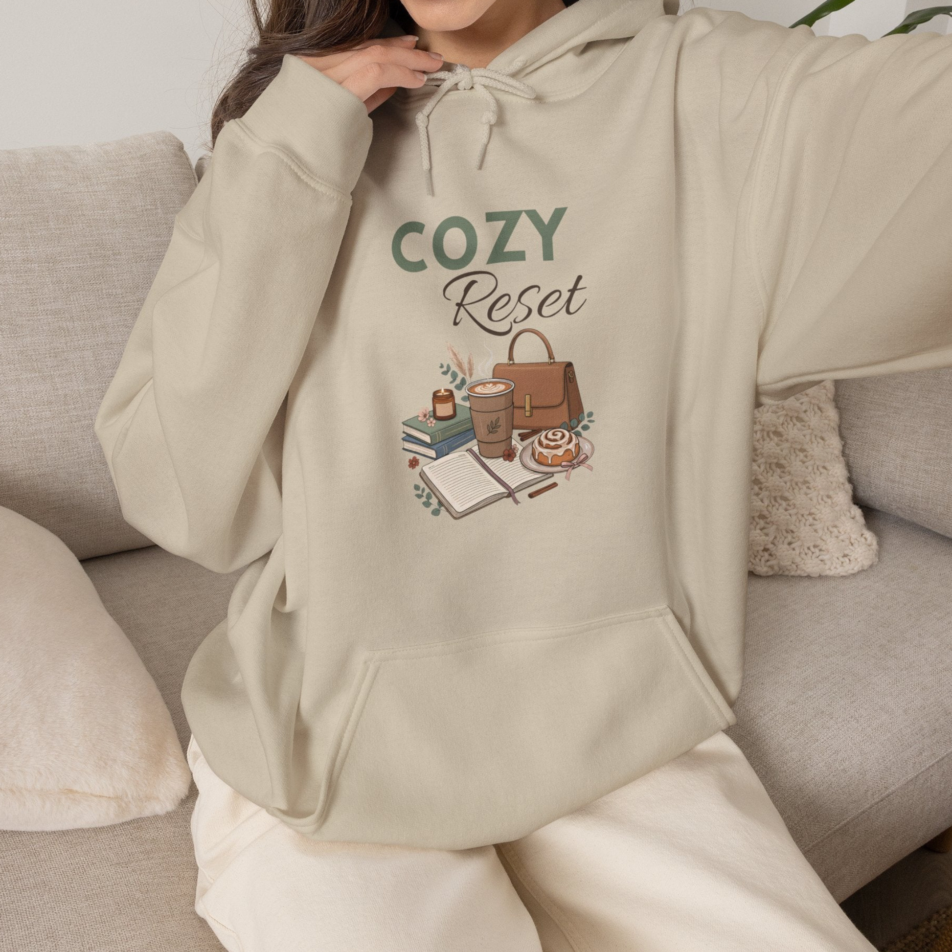 A woman is wearing a sand-colored hooded sweatshirt with 'COZY Reset' on the front and is sitting on the couch.