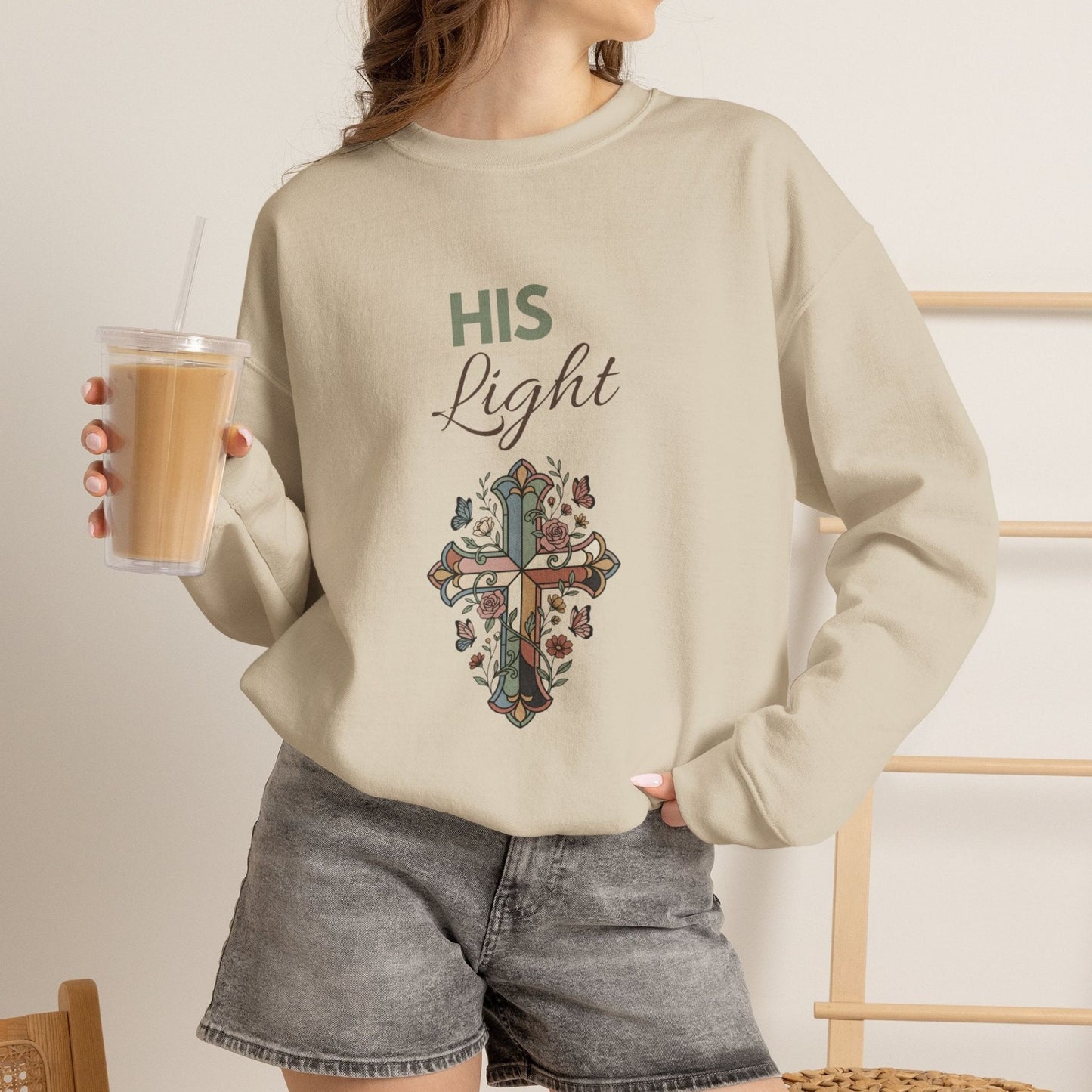 A woman is wearing a sand-colored sweatshirt with 'His Life' on the front and is holding an iced coffee.