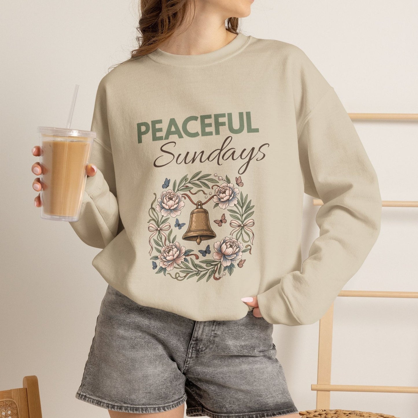 A woman is wearing a sand-colored sweatshirt with 'PEACEFUL Sundays' on the front and is holding an iced coffee.
