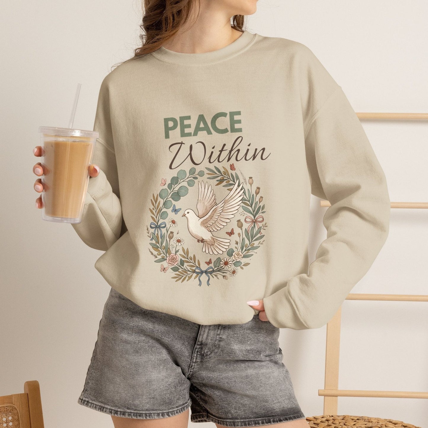 A woman is wearing a sand-colored sweatshirt with 'PEACE Within' on the front and holding an iced coffee.