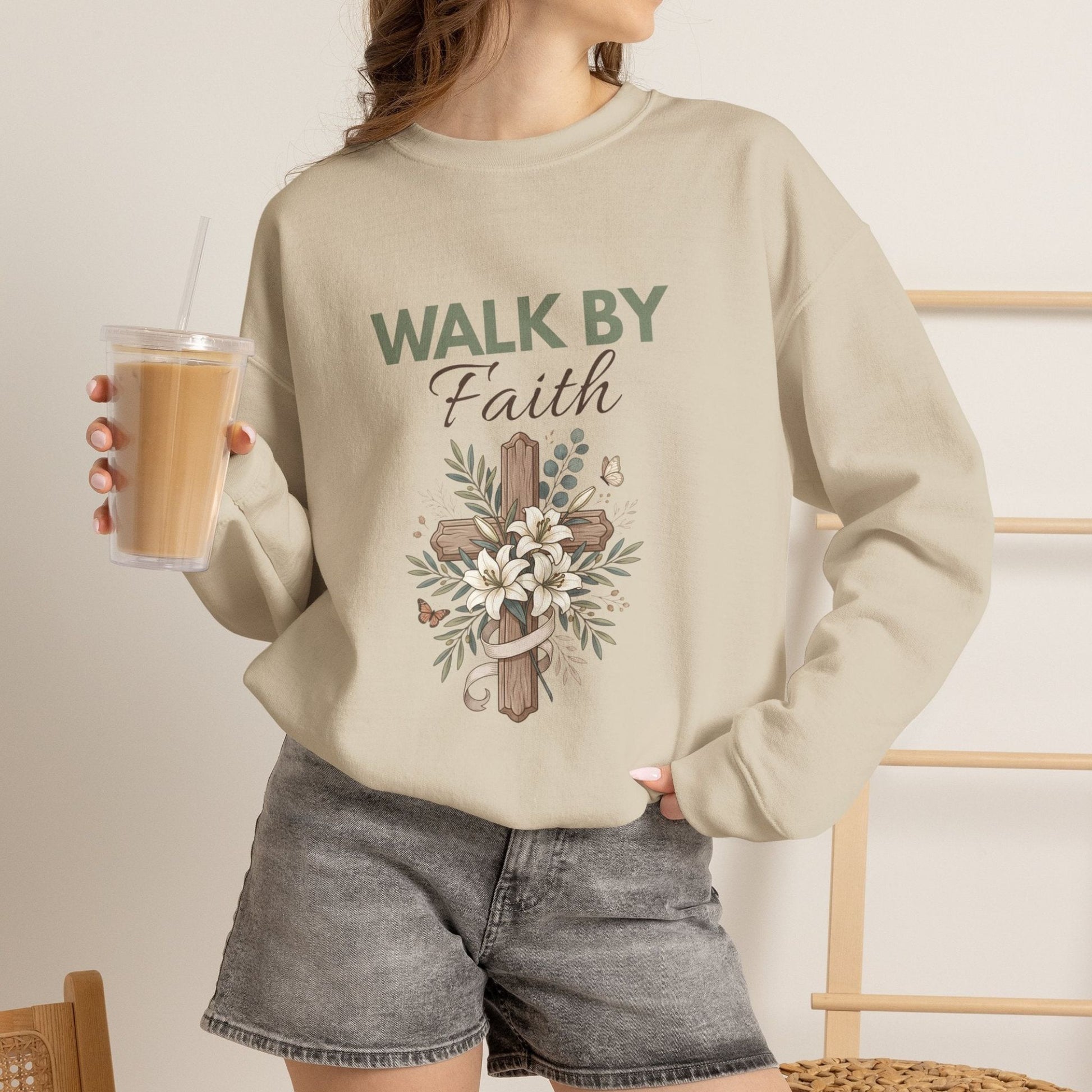 A woman is holding an ice coffee while wearing a sand colored crewneck sweatshirt with 'WALK BY Faith' on the front.