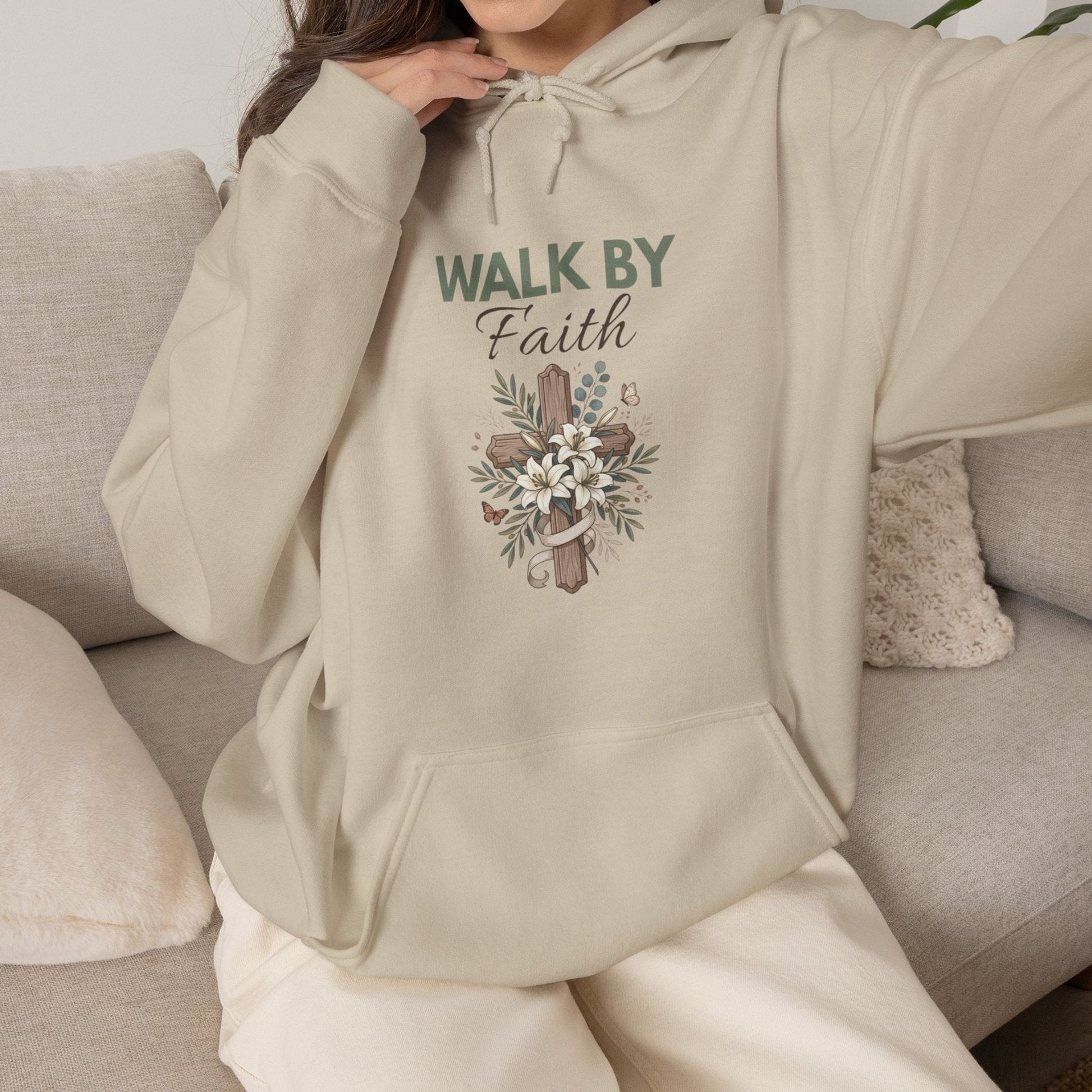 A woman is sitting on a couch while wearing a sand colored hooded sweatshirt with 'WALK BY Faith' on the front.