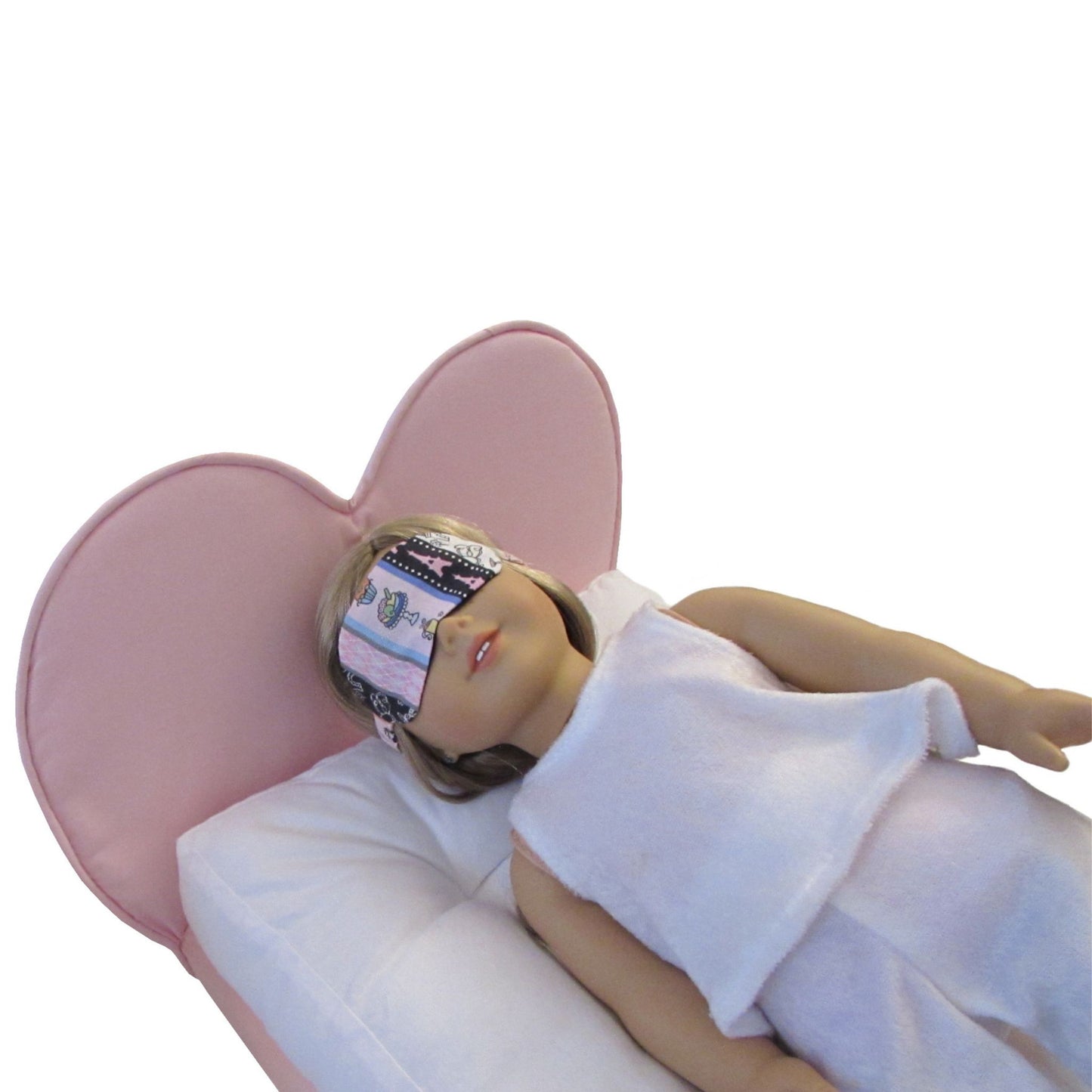 An 18-inch doll wearing a striped Paris print sleep mask while lying on a pink heart doll bed.