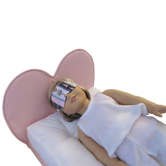 An 18-inch doll wearing a striped Paris print sleep mask while lying on a pink heart doll bed.