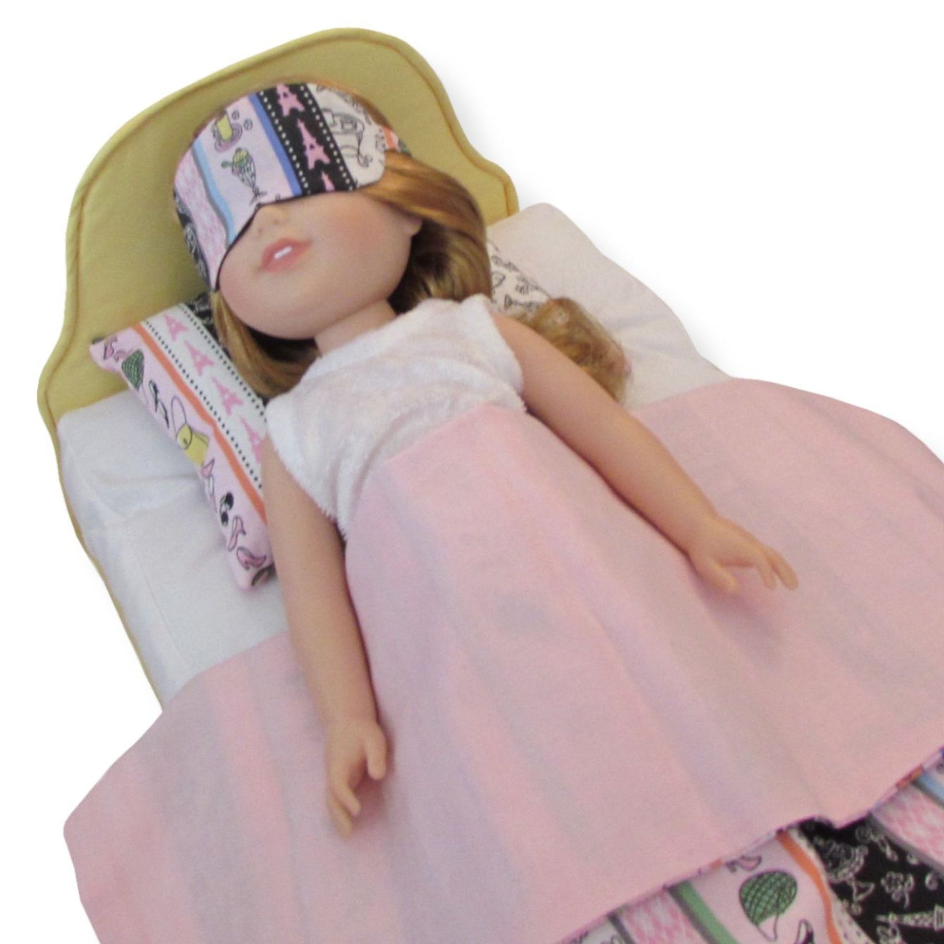 A 14.5-inch doll wearing a striped Paris print sleep mask while lying in a yellow doll bed.