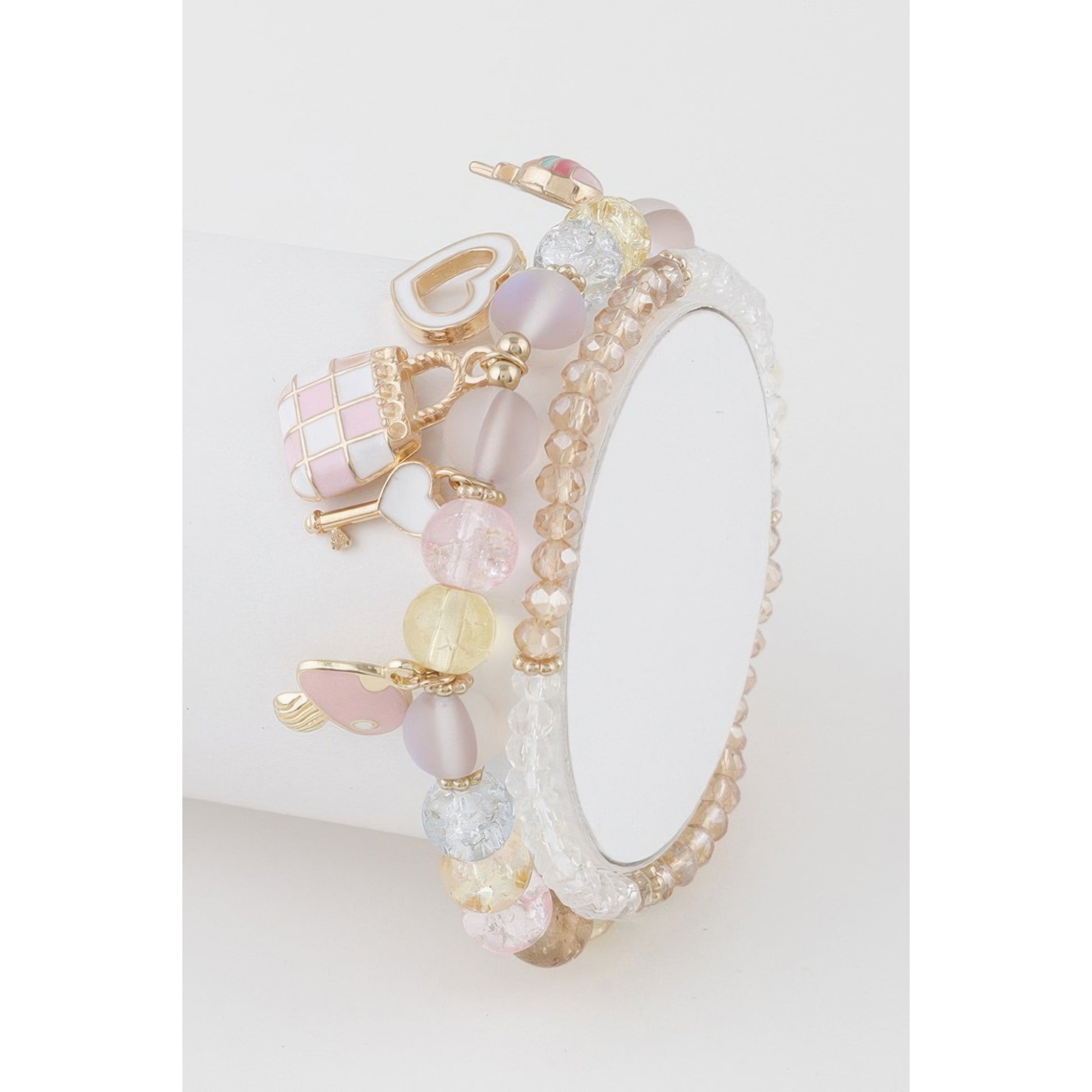 A delicate charm bracelet with pastel pink, lavender, and gold elements, displayed on a white cylindrical stand against a clean white background.