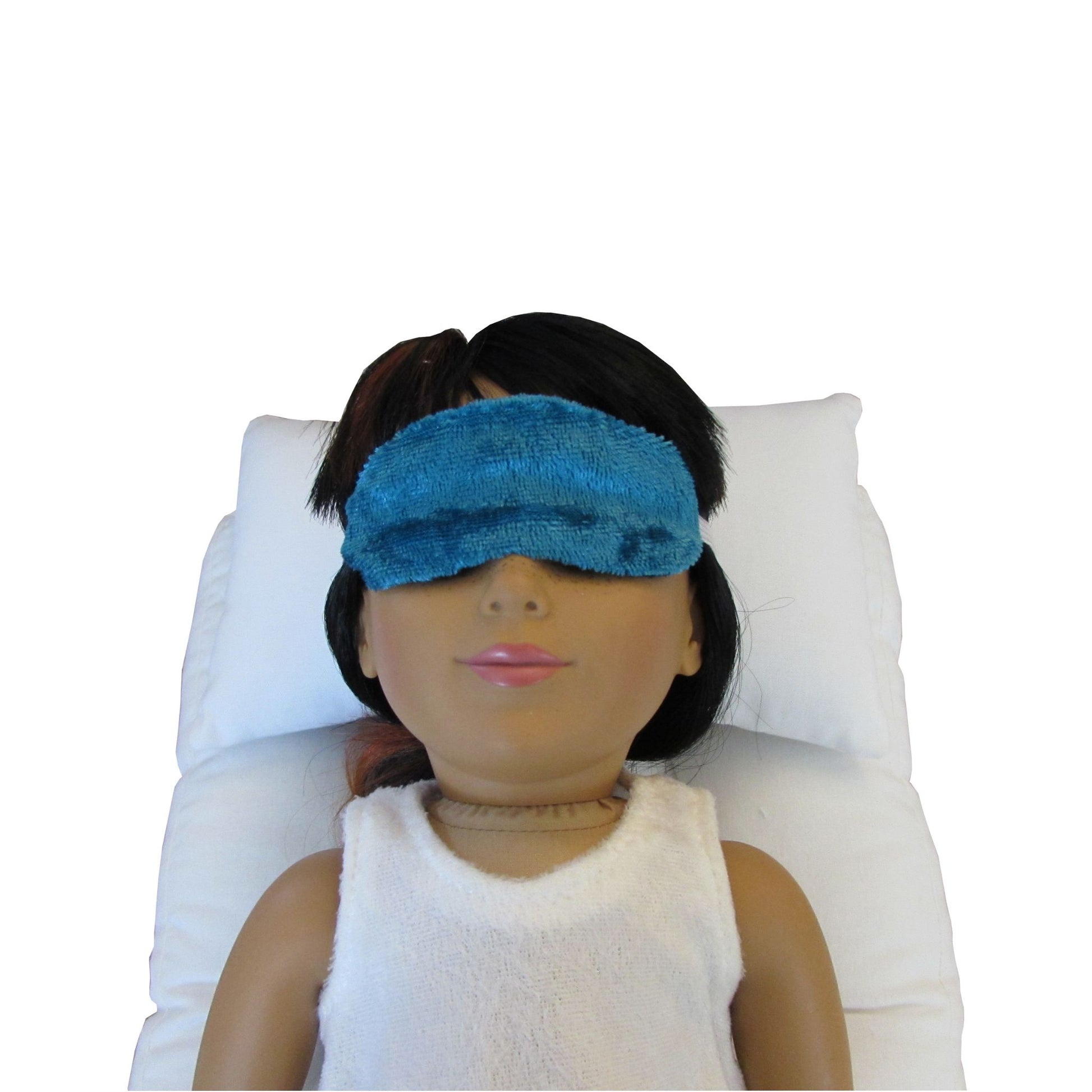 An 18-inch doll is wearing a turquoise sleep mask while lying on a white doll pillow and a white doll mattress.