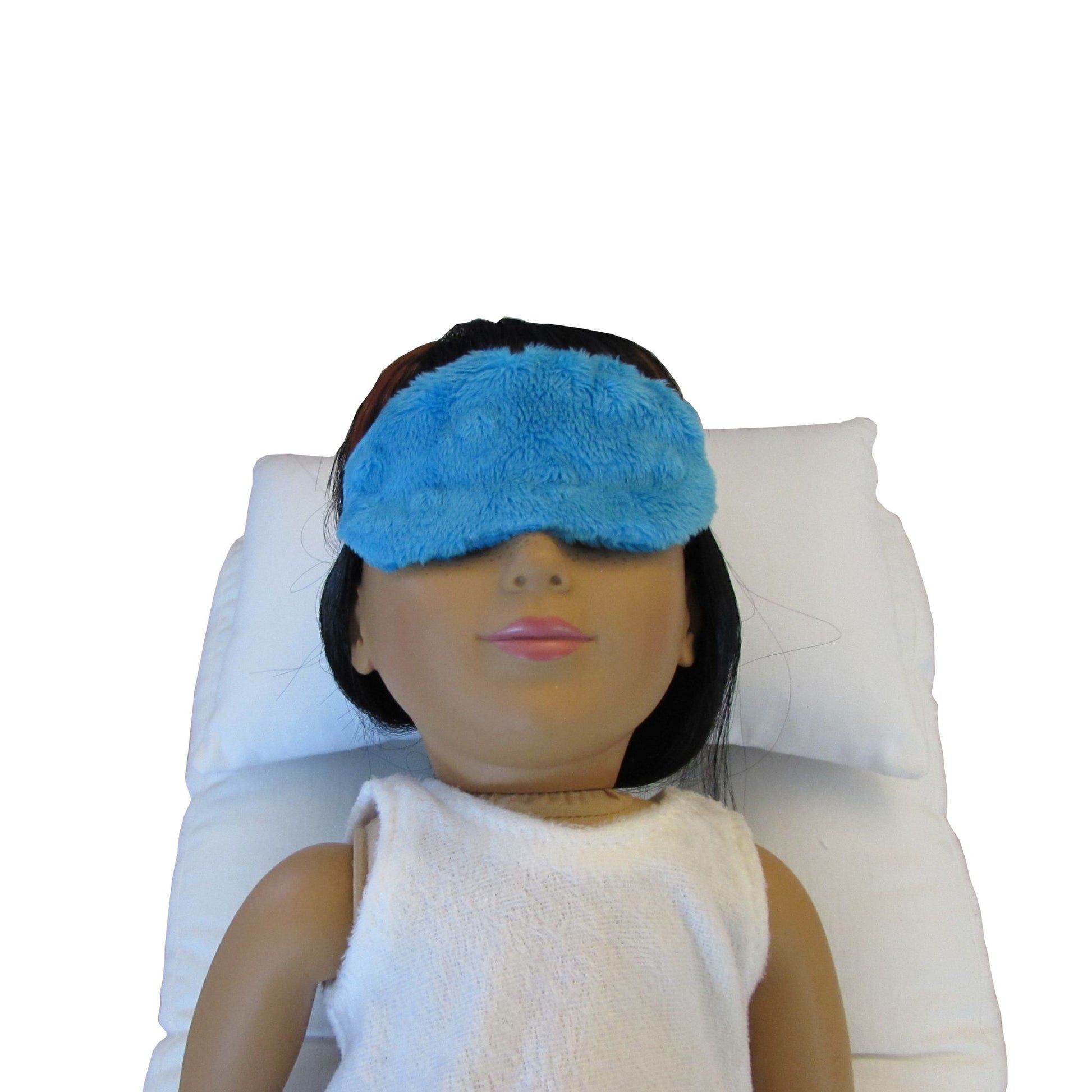 An 18-inch doll is wearing a turquoise minky sleep mask while lying on a white doll pillow and a white doll mattress.
