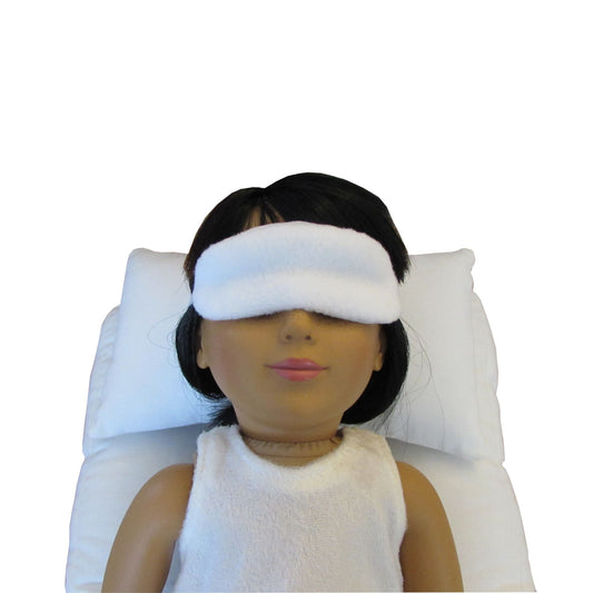 An 18-inch doll is wearing a white anti-pill fleece sleep mask while lying on a white doll pillow and a white doll mattress.