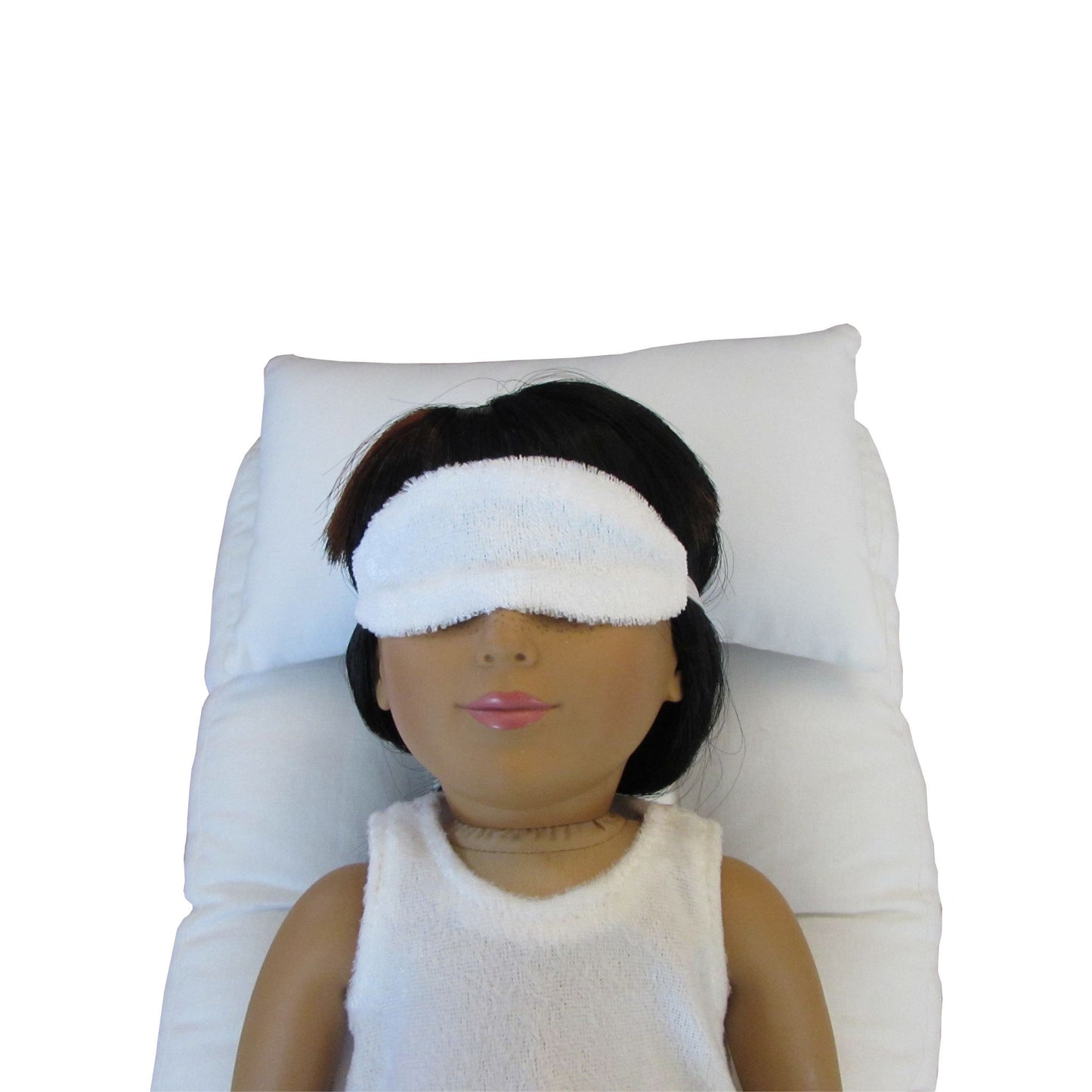 An 18-inch doll wearing a white panne velvet sleep mask while lying on a white doll pillow and a white mattress.