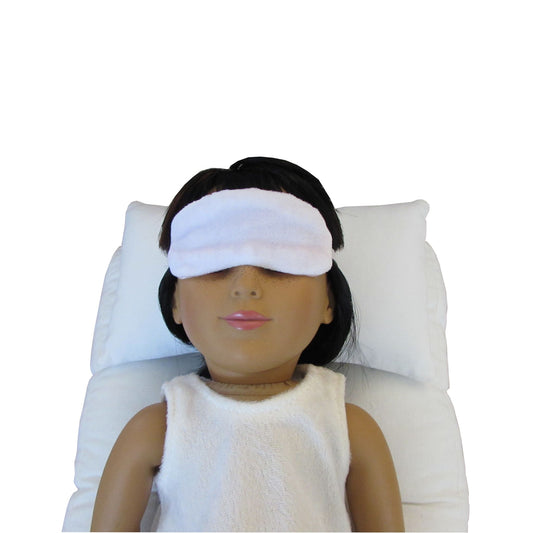 An 18-inch doll is wearing a white stretch velvet sleep mask while lying on a white doll pillow and a white doll mattress.