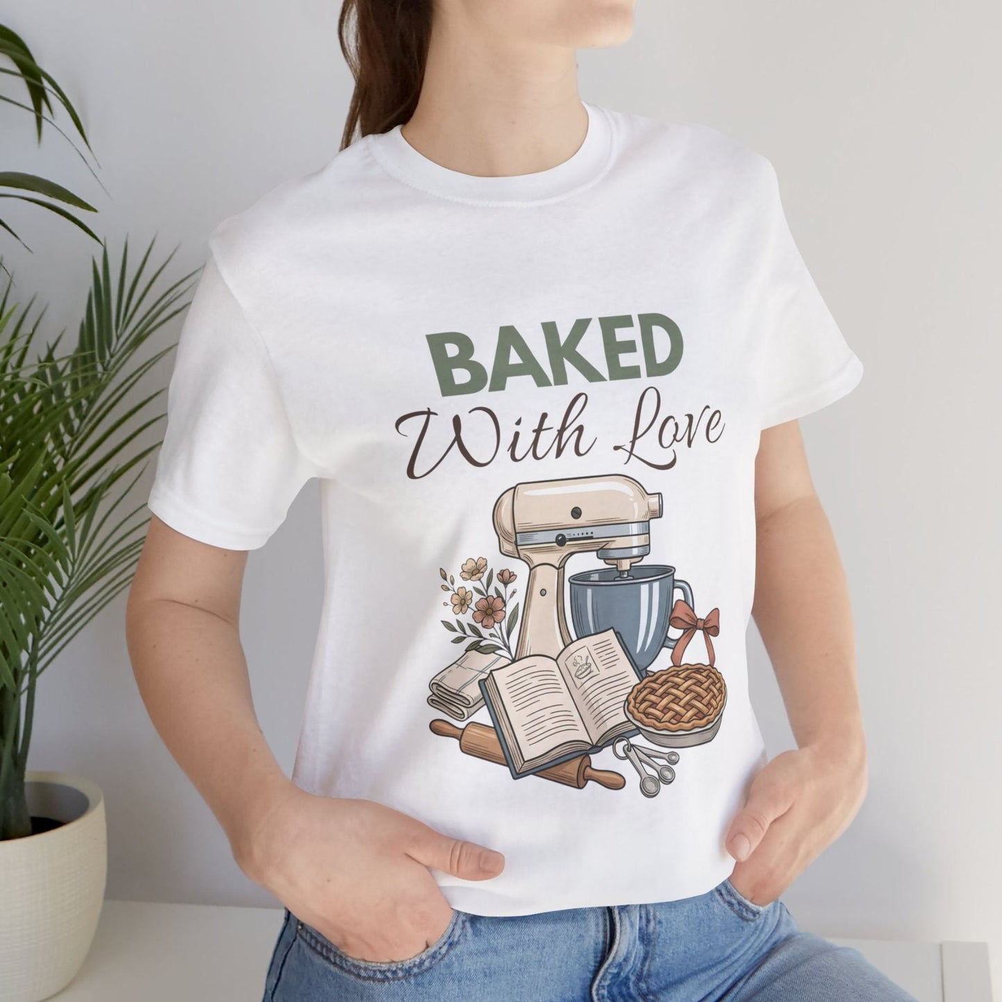 A woman wearing a white t-shirt with "BAKED With Love' on the front standing by a plant.