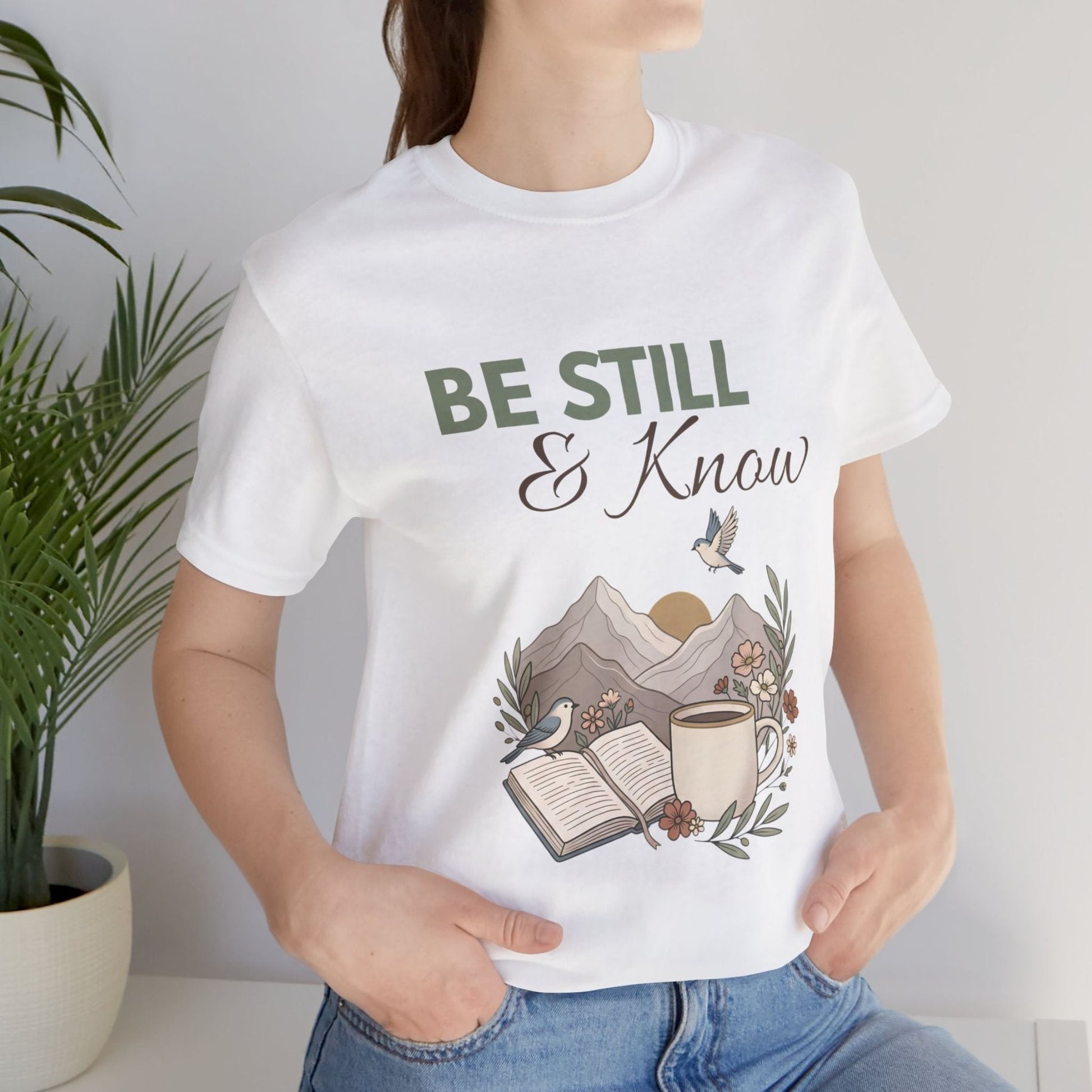 Person wearing a white t-shirt with 'Be Still & Know' design, standing next to a plant.