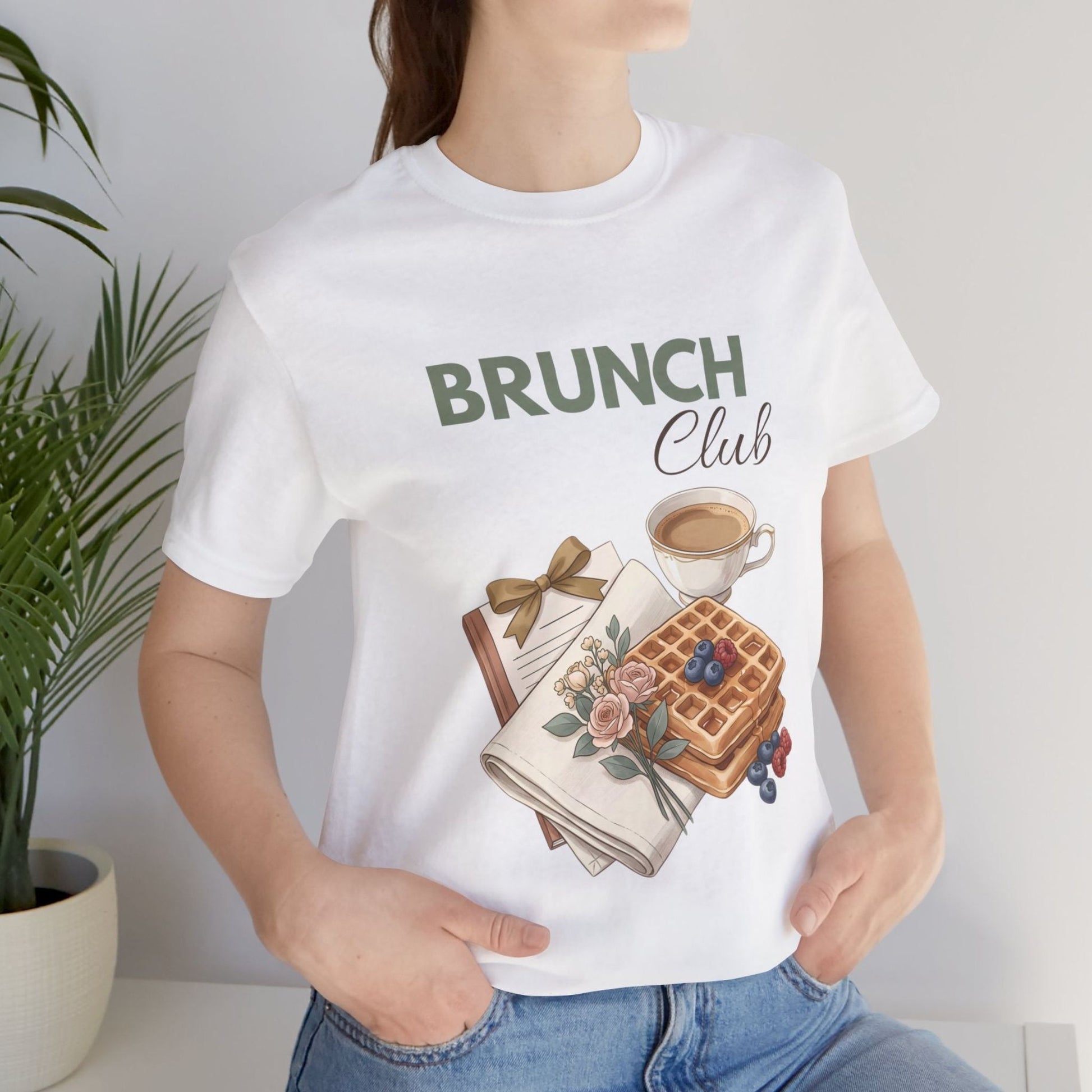 A woman is wearing a white t-shirt with 'BRUNCH Club' on the front while standing by a plant.