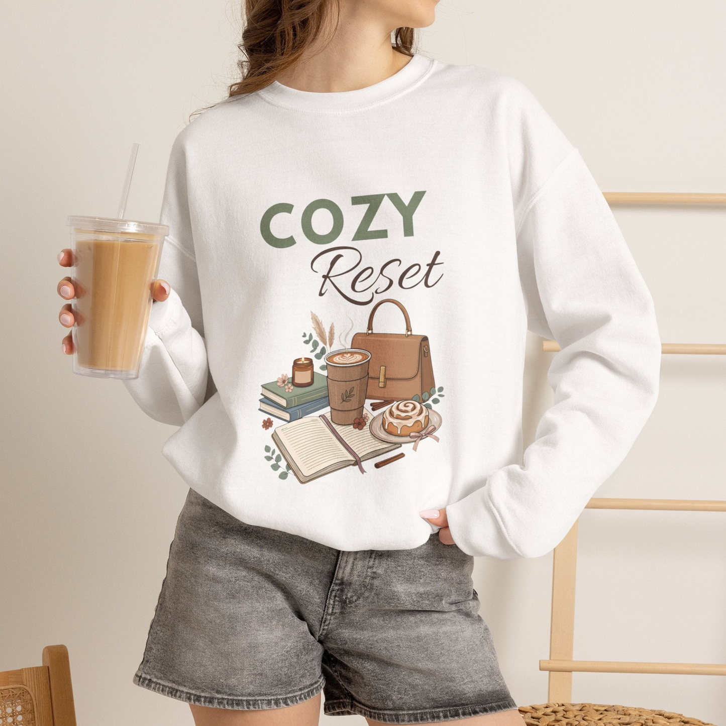 A woman is wearing a white crewneck sweatshirt with 'COZY Reset' on the front and is holding an iced coffee.