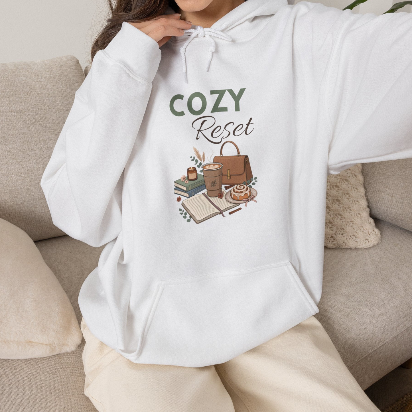 A woman is wearing a white hooded sweatshirt with 'COZY Reset' on the front and is sitting on a couch.