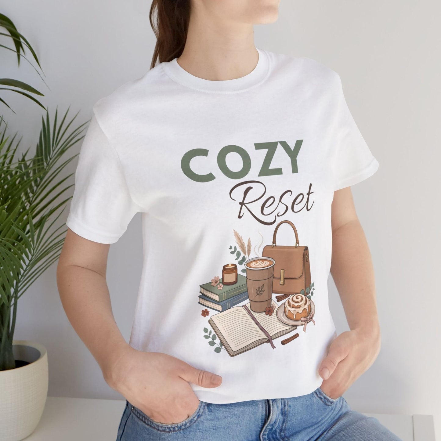 A woman is standing by a plant while wearing a white t-shirt with 'COZY Reset' on the front.