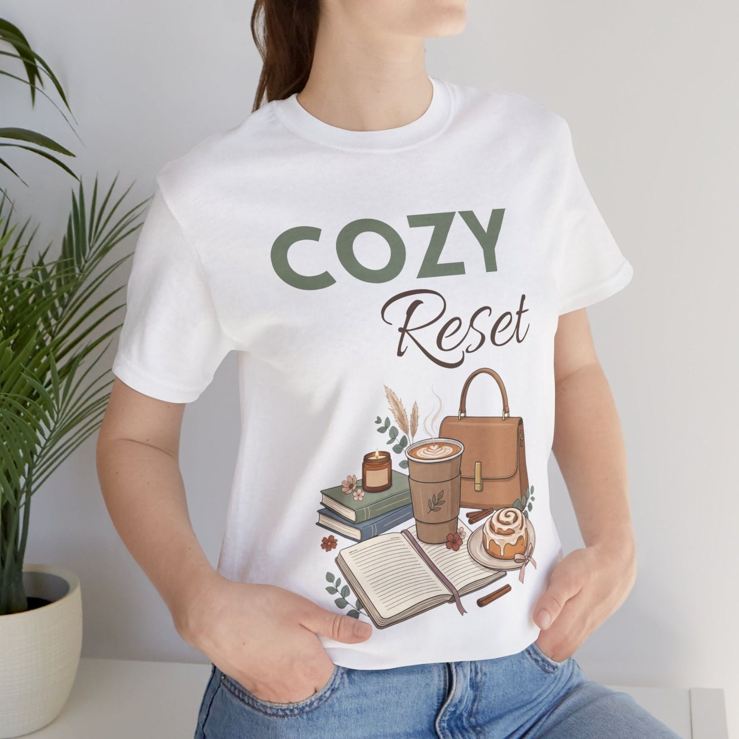 A woman is wearing a t-shirt with 'Cozy Reset' on front while sitting by a plant.