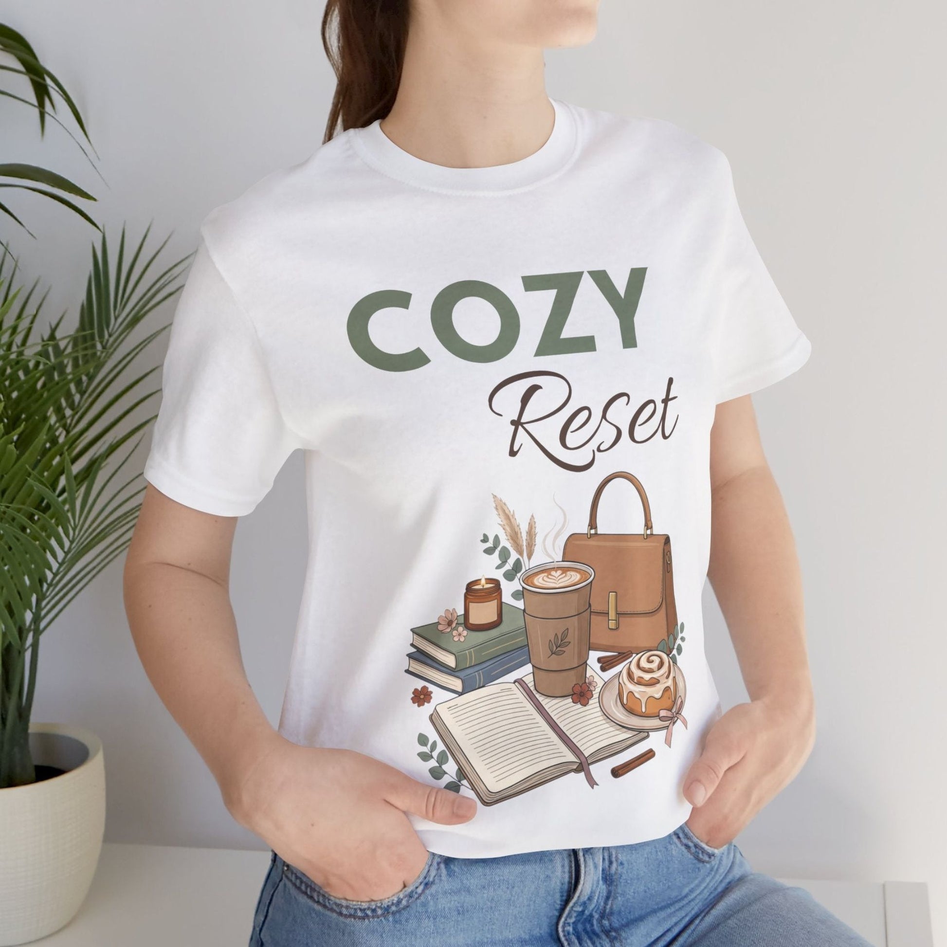 A woman is wearing a t-shirt with 'Cozy Reset' on front while sitting by a plant.