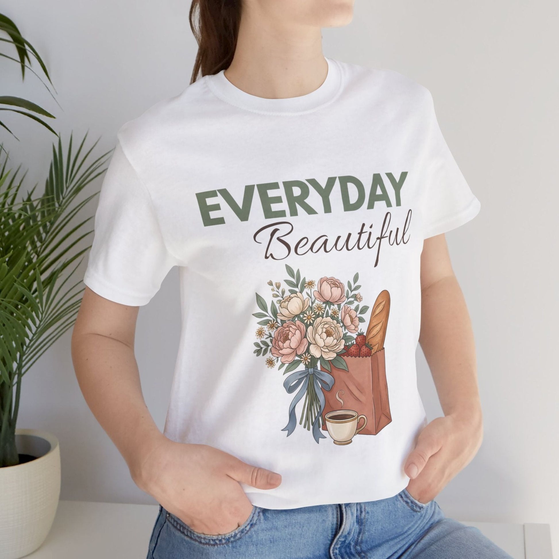 A woman wearing a white t-shirt with "EVERYDAY Beautiful' on the front standing by a plant.
