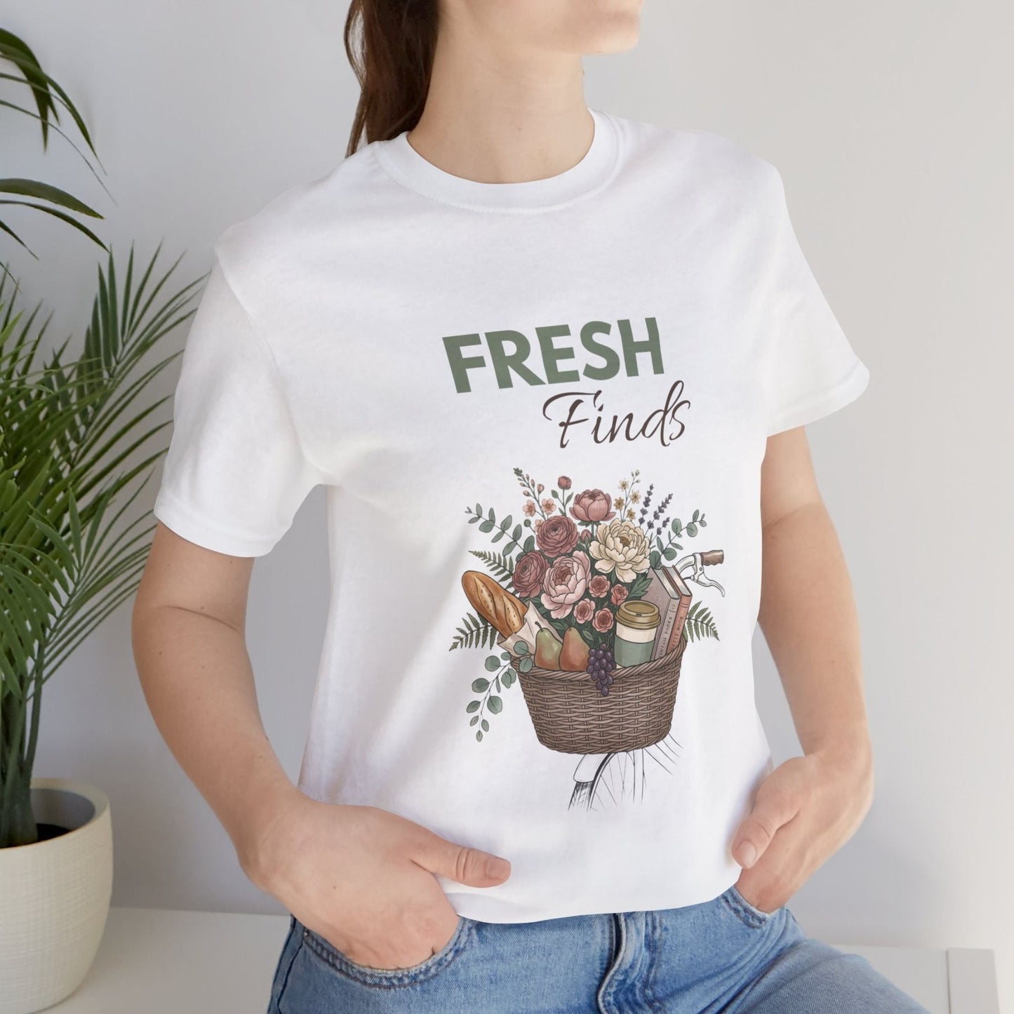 A woman standing by a plant while wearing a white t-shirt with 'FRESH Finds' on the front.