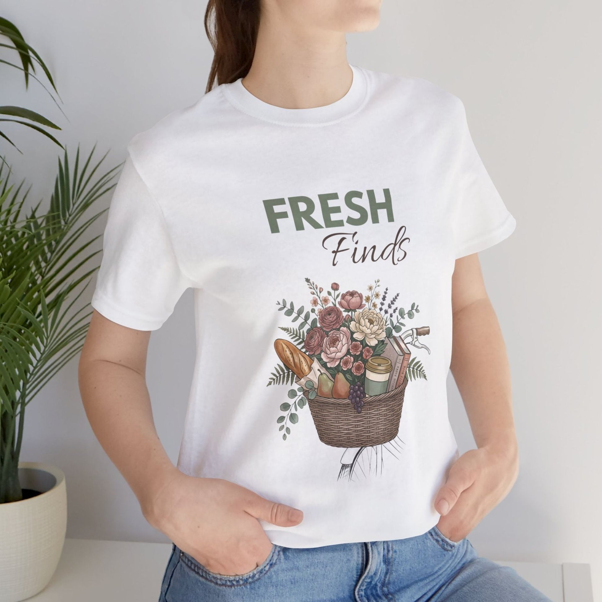 A woman standing by a plant while wearing a white t-shirt with 'FRESH Finds' on the front.