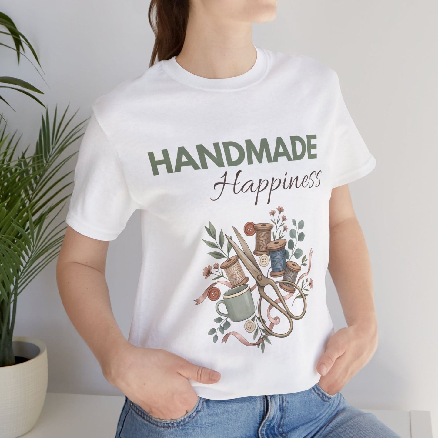 A woman wearing a white t-shirt with 'Handmade Happiness' on the front while standing by a plant.
