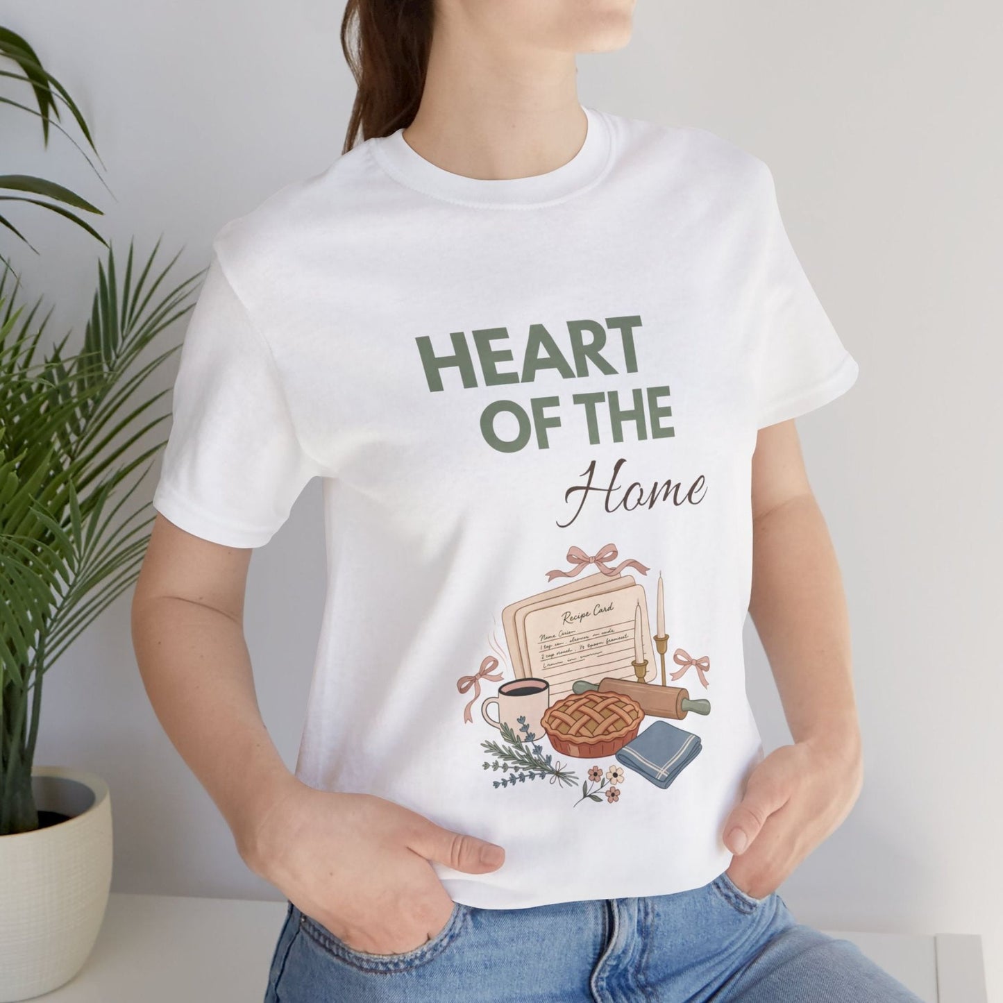 A woman is standing by a plant while wearing a 'HEART OF THE Home' t-shirt.