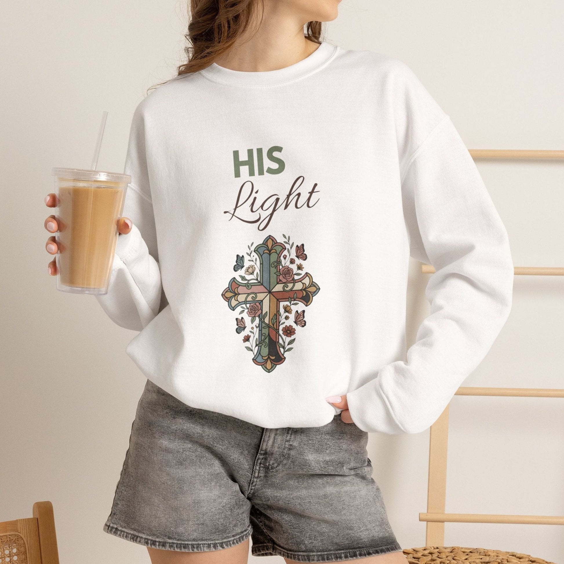 A woman is wearing a white sweatshirt with 'His Light' on the front and is holding an iced coffee.