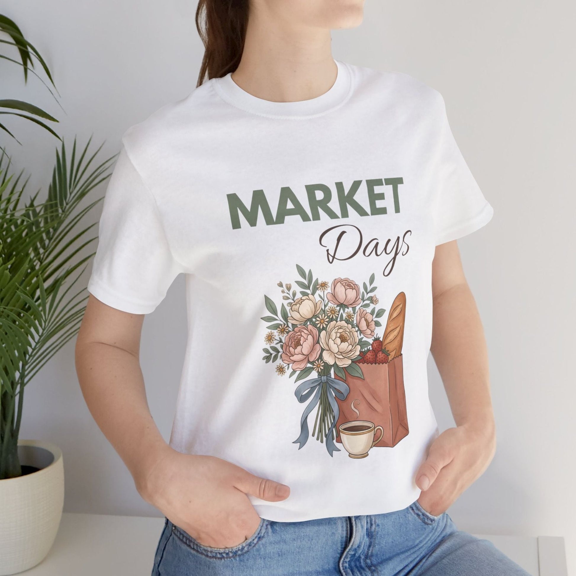 A woman wearing a white t-shirt with 'Market Days' on the front standing by a plant.