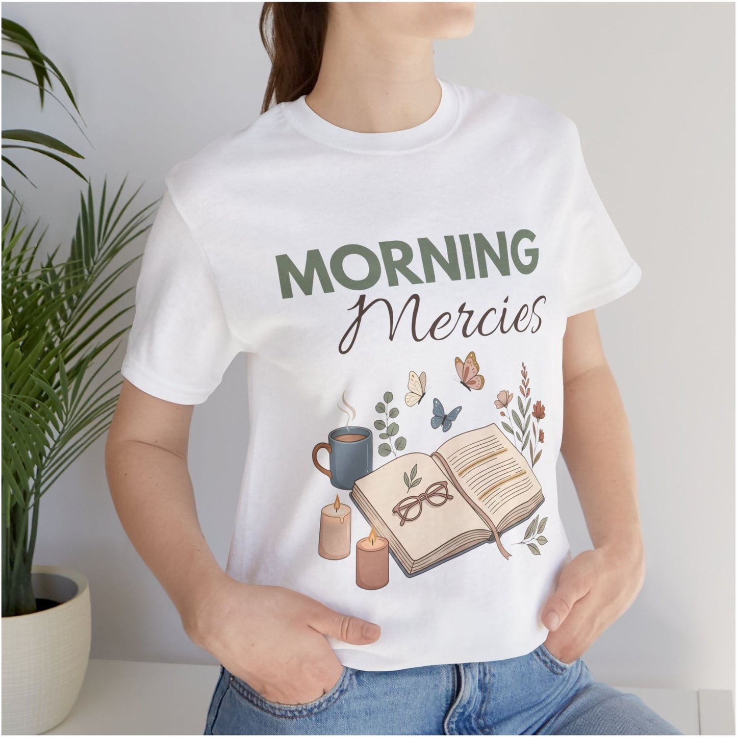 Person wearing a white t-shirt with 'Morning Mercies' design, including a book, coffee cup, and candles.